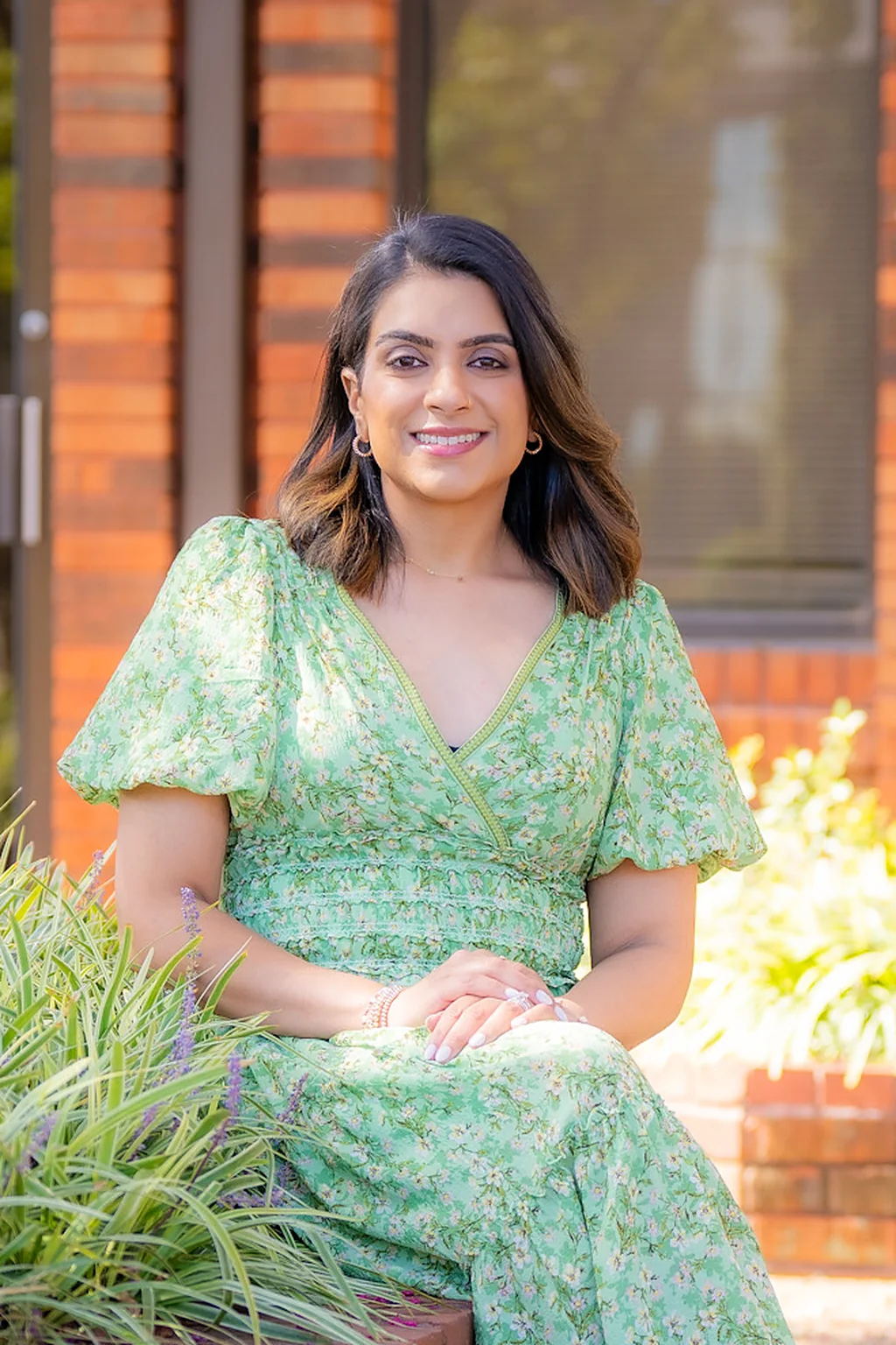 Priya Patel, PA-C, smiling, in a green floral dress, seated by a planter outside the office